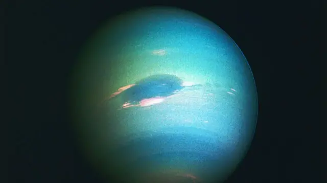Neptune is seen illuminated as an almost-full sphere, with different shades of greenish-blue bands across its surface and a large deep blue oval area in the middle.