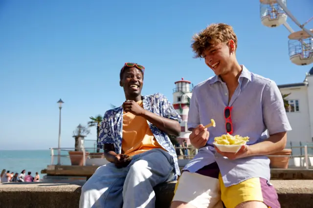 Dos hombres en la playa riendo mientras uno sostiene algo de comida.