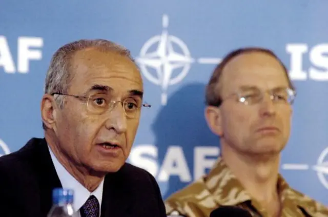 Takım elbiseli bir erkek, yanında askeri üniformalı başka bir erkek. Arkadaki mavi panoda NATO ve ISAF logosu var.