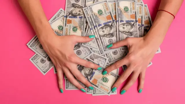 Turkuaz ojeli tırnaklara sahip iki el, büyük bir yığın 100 dolarlık banknotun üzerine yayılmış, parayı örtüyor ve görünüşe göre topluyor. Sahne, parlak pembe bir arka plan üzerinde yakın çekim bir görüntüdür.