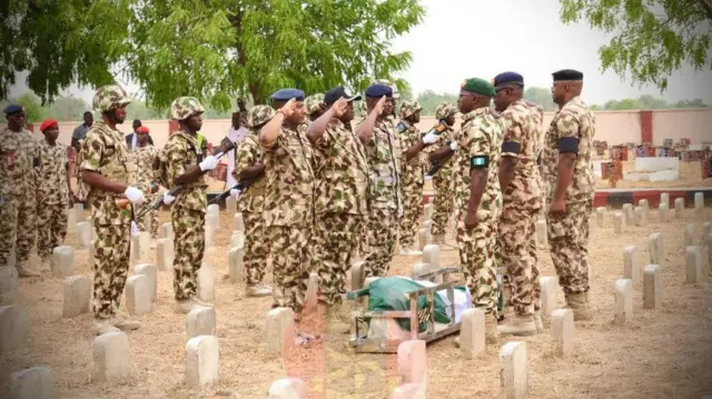 Nigerian Army Officers stand for cemetry dey salute as deadibody of one of di sojas wey kpai dey for ground, dem wrap am wit Nigeria flag of green and white.