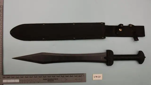 Uma espada colocada sobre um lençol de cor clara ao lado de um estojo usado para guardar a arma. Há uma régua ao lado da espada
