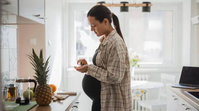 Mulher grávida, em pé, parada em frente ao balcão da cozinha, retirando uma cápsula de um frasco