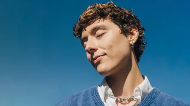 A man closes his eyes while the sun shines on his face. He has short brown curly hair and is wearing a white shirt and blue jumper, with a silver necklace. Behind him is the bright blue sky.