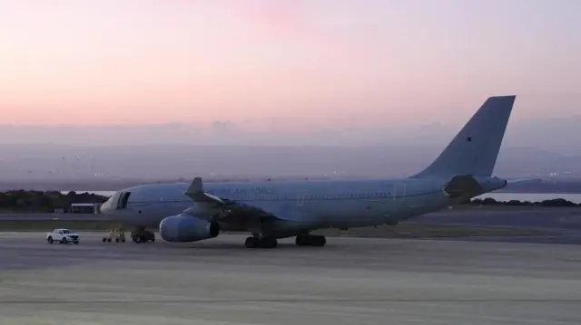 A Voyager aircraft - which resembles a passenger airliner and is grey with 'Royal Air Force' written on the side in a darker grey - sits on the runway at RAF Akrotiri. The sun is setting in the background.