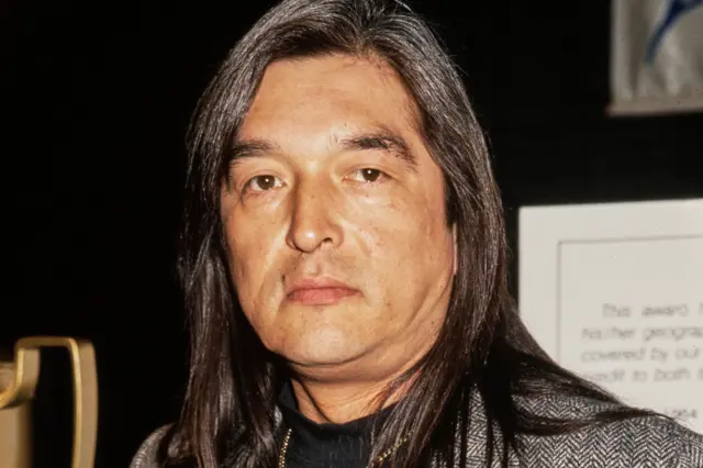 Graham Greene has long straight hair and is wearing a patterned blazer over a dark top.