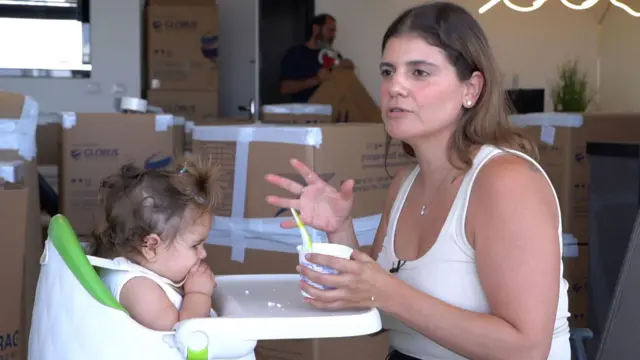 Krem rengi üst giyinmiş,, kahverengi saçlı bir kadın bebek masasının karşısında konuşurken elinde mama kabı ve kaşığı tutuyor Bebek de masada bir şeyler yiyor. Arka planda çok sayıda kutu var. 