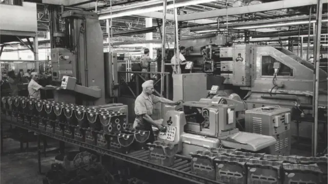 A imagem em preto e branco mostra uma linha de produção industrial dentro de uma fábrica ampla, com operários trabalhando em diferentes máquinas e equipamentos metálicos. Em primeiro plano, peças mecânicas alinhadas sobre uma esteira passam por processos de usinagem enquanto trabalhadores operam painéis de controle e acompanham a produção. O ambiente é iluminado por estruturas industriais no teto e tem aparência de registro histórico de uma fábrica automotiva ou metalúrgica.