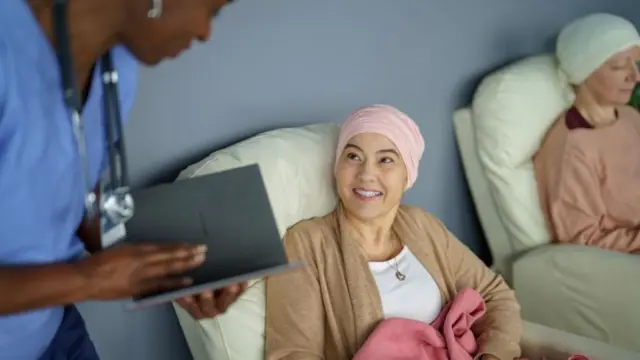 Paciente de câncer, com toalha na cabeça, sentada em uma poltrona, conversa com profissional de saúde