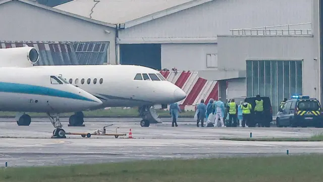 PPE-clad officials wait near a jet on the tarmac at an airport.