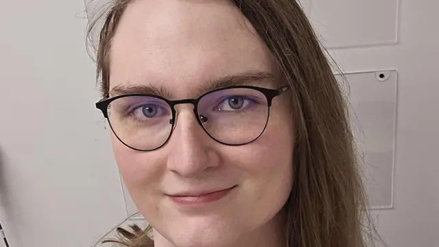 A close-up image of a face with long hair and glasses, smiling slightly directly at the camera