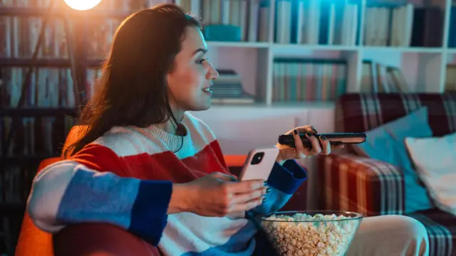 A woman sitting on the couch alone, watching TV and using her phone.