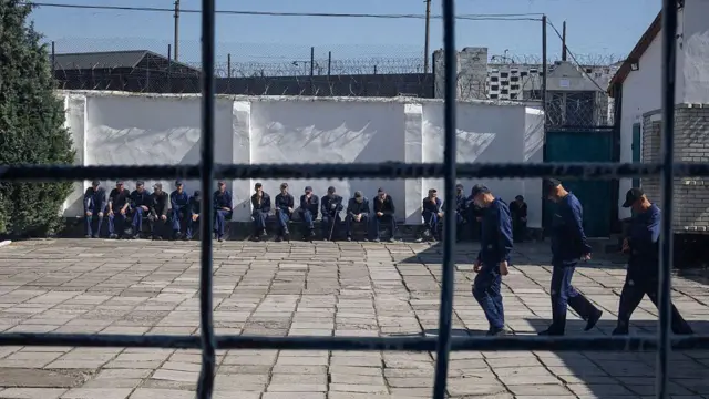 Залитый солнечным светом тюремный двор, по которому шагает шеренга заключенных, а ряд других заключенных, в тюремной одежде, сидит на скамейке на заднем плане.