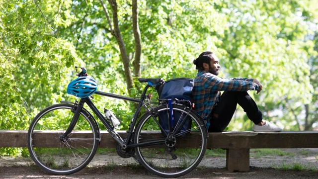 Um homemlenny slotcabelos compridos, presoslenny slotum coque, está sentado ao ladolenny slotsua bicicleta. Ele veste uma camisa xadrez, calças pretas e tênis brancos.