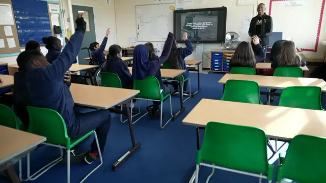 Niños con uniformes azul marino levantan las manos desde detrás de escritorios de plástico y sillas de plástico verde mientras un hombre permanece frente a una pantalla de televisión.