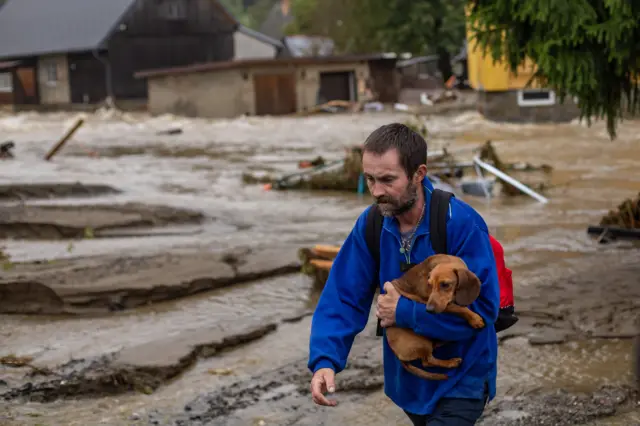 Homem de casaco azul carrega seu cachorro, com rua alagada vista ao fundo