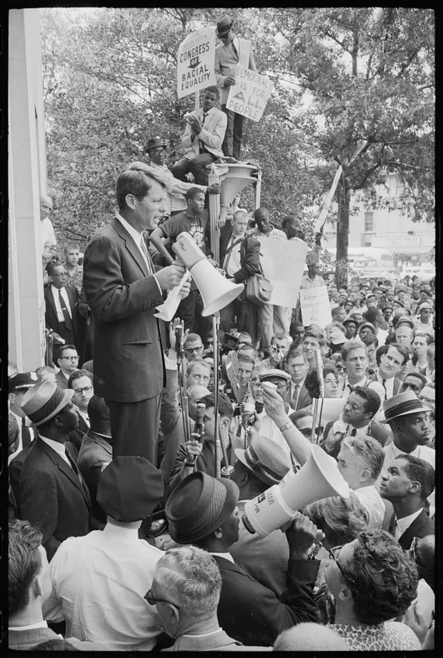 Robert F. Kennedy discursando com megafone, rodeado por multidão