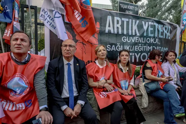 Mahmut Tanal eğitim-iş sendikasından eğitimcilerle bir duvarın üstünde oturuyor. arkada "artık yeter, güvenli okul, sağlıklı eğitim ortamı istiyoruz" yazılı bir pankart var. 