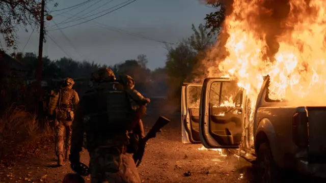 Trzech uzbrojonych żołnierzy w mundurach sprawdza nocą płonący pojazd. Z otwartych drzwi ciężarówki wydobywają się płomienie.