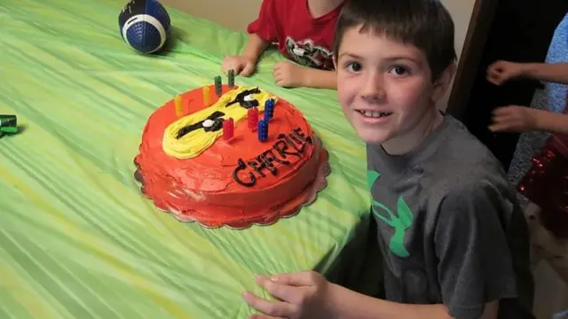 Uma criança sorrindo para foto. Na mesa tem um bolo laranja com o nome dele e velas