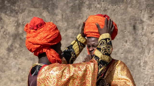 A man in a colourful robe and turban helps another adjust his outfit.