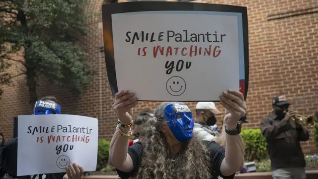 Manifestantes com máscaras azuis mostram cartazes com os dizeres: "Sorria, a Palantir está observando você"
