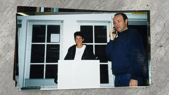 Kısa saçlı bir kadın, Ghislaine Maxwell, gülümsüyor ve beyaz bir kürsünün arkasında duruyor. Solunda bir erkek, Kevin Spacey, sağ kulağında cep telefonuyla duruyor. Lacivert bir gömlek ve mavi kot pantolon giyiyor.