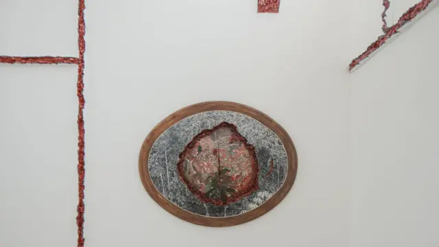 Em uma parede branca marcada por fissuras avermelhadas que parecem atravessar a arquitetura, uma obra oval emoldurada em madeira revela uma paisagem vegetal parcialmente rasgada por uma abertura orgânica e visceral.