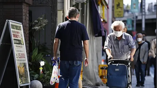 Idoso caminhando na calçada com caminhador e máscara