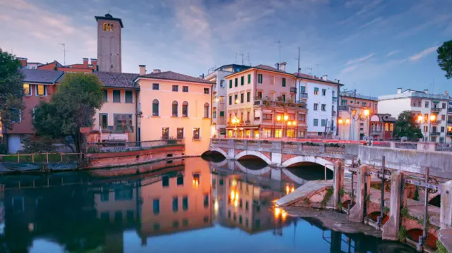 Clădiri vechi colorate și un turn se văd reflectate în suprafața de apă a unui canal din Treviso. 