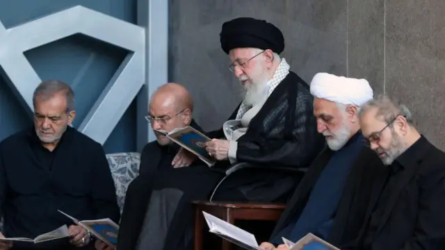 المرشد الأعلى الإيراني علي خامنئي، يرتدي جلباباً رمادي اللون وعباءة سوداء وعمامة سوداء بينما يقرأ القرأن ويتوسط الرئيس الإيراني مسعود بزشكيان ورئيس البرلمان محمد باقر قاليباف.