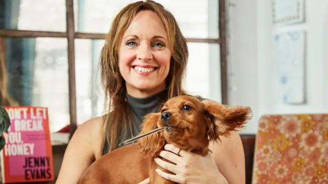 Дженні Еванс посміхається в камеру, вона тримає на руках коричневе цуценя з ручкою в роті. Позаду Дженні лежить книга з написом "Не дозволяй цьому зламати тебе, люба" Дженні Еванс.