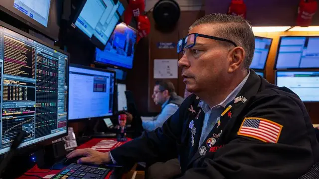 米ニューヨーク証券取引所（NYSE）の男性トレーダーが、眼鏡を額に乗せ、モニターを見ている。ダークカラーのジャケットの左腕に星条旗のワッペンがついている