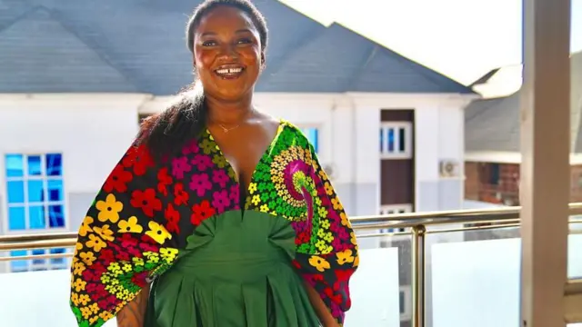 Susan wear colourful dress. She smile give camera as she stand for balcony. You fit see anoda house behind her.