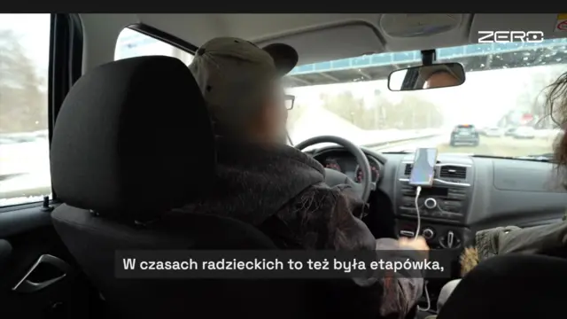 Салон автомобиля, снятый с заднего сиденья. На переднем плане – спинка кресла водителя и сам водитель в кепке и прямоугольных очках. Его лицо неаккуратно замазано, видны скула и висок. Через лобовое стекло просматривается зимняя дорога, а справа от водителя – пассажирка на переднем сиденье. Внизу субтитр: «В советское время это тоже был этапный пункт».