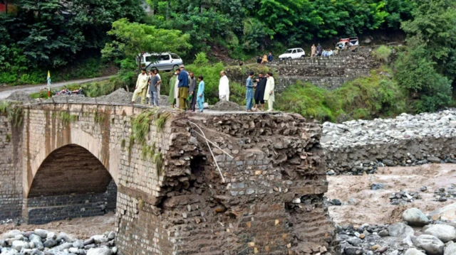 বন্যার পানির তোড়ে ভেঙে যাওয়া একটি ব্রিজের ওপর দাঁড়িয়ে আছেন কয়েকজন লোক