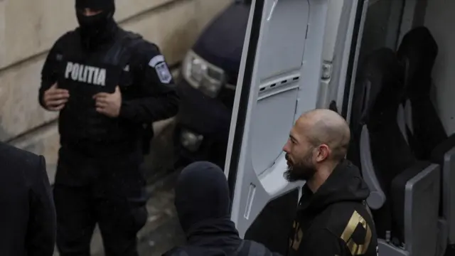Andrew Tate eskotowany przez rumuńską policję po zatrzymaniu w Bukareszcie. Tate stoi w otoczeniu trójki funkcjonariuszy przed otwartą białą furgonetką. 12 marzec 2024 r.
