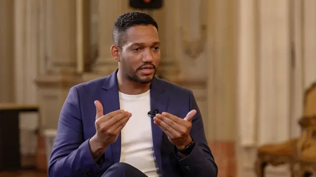 Rick Azevedo gesticulando durante entrevista à BBC. Ele veste terno azul e camiseta branca, e usa um cavanhaque e cabelo curto, raspado dos lados