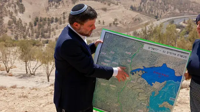 وزير المالية الإسرائيلي اليميني المتطرف، بتسلئيل سموتريتش، يعرض خريطةً لمنطقة قرب مستوطنة معاليه أدوميم، في 14 أغسطس/آب 2025، وأعلن حينها عن مشروع لبناء بؤرة استيطانية في المنطقة المعروفة باسم "E1" (إي وان)، ويظهر في الصورة سموتريتش مرتدياً سترة زرقاء داكنة وقميصاً أبيض و قلنسوة وأمامه خريطة.