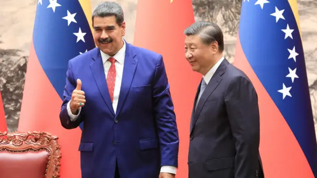 Nicolás Maduro, ao lado de Xi Jinping, com bandeiras da China e da Venezuela atrás deles. Ambos vestem ternos escuros e gravata e estão sorrindo. Maduro faz sinal de positivo com o polegar.