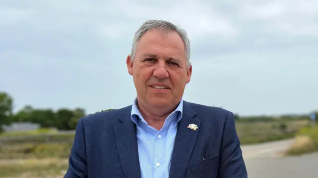 El agente Andy Jehan, un hombre de pelo corto y canoso, está bien afeitado y viste una chaqueta azul marino sobre una camisa azul cielo con botones blancos. Lleva el botón superior desabrochado. Lleva una placa en la solapa izquierda. Detrás de él, desenfocada, se ve una carretera y campos verdes.