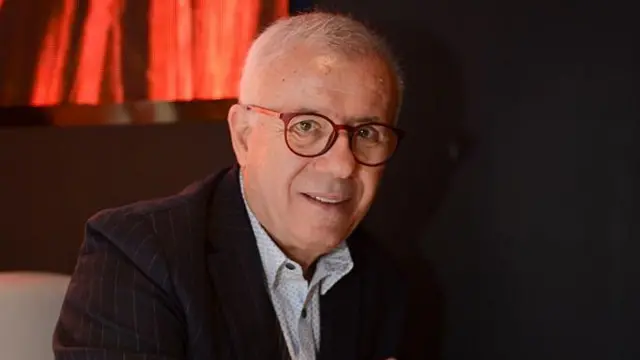 Beyaz seyrek saçlı bir erkek kravatsız takımı ile oturarak poz veriyor. Gözlükleri yuvarlak.