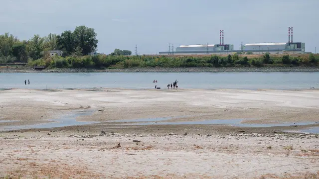 9 августа 2026 года люди на берегу Дуная, уровень воды в котором крайне низкий, недалеко от города Пакш (Венгрия); на заднем плане видна Пакшская атомная электростанция