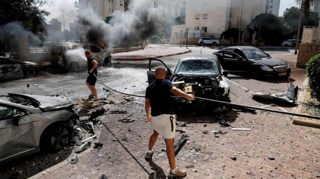 Pessoas trabalham para apagar um incêndio que envolve uma van, enquanto foguetes são lançados da Faixa de Gaza