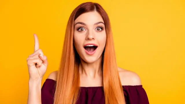Retrato fotográfico en primer plano de una mujer joven con cabello largo rubio fresa, con una blusa vinotinto que deja ver los hombro, con la boca abierta, mirando a la cámara y sosteniendo el dedo índice levantado sobre un fondo brillante aislado.