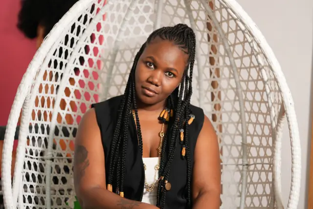 Chiamaka Richards yi uwe ọcha n'ime ma yikwasị ya uwe ojii buru isi a kpara akpa, nyara ọlaolu ma nọrọ ọdụ n'oche a kpara akpa na-acha ọcha.