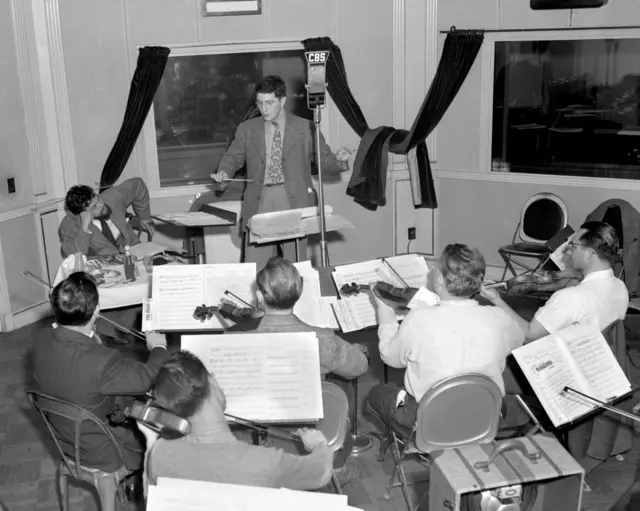 Várias pessoas com violinos tocando e um homem no centro regendo