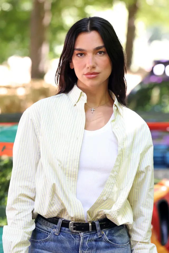 Portret Dua Lipy w swobodnym stroju: niebieskie dżinsy z czarnym paskiem, biały T-shirt i zielona koszula. Ma naszyjnik z małym krzyżykiem i rozpuszczone ciemne włosy.