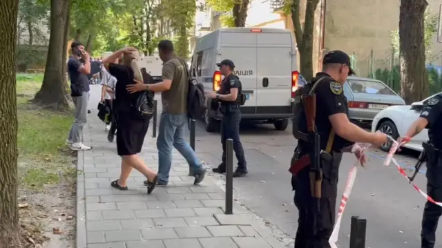 Поліція огороджує місце злочину стрічкою. Жінка у центрі кадру йде, схопившись за голову, її за спину притримує чоловік