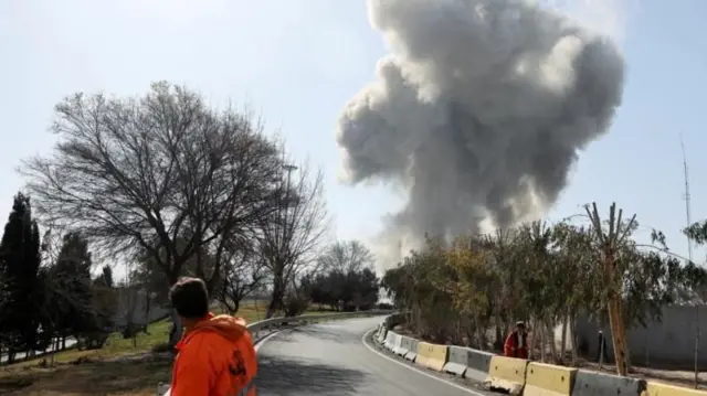 İleride duman yükseliyor, bir adam arkası dönük bir yoldan karşıya geçiyor. 